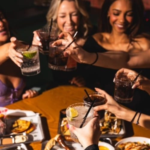 Group of smiling women toasting cocktails at a dinner table with shared appetizers
