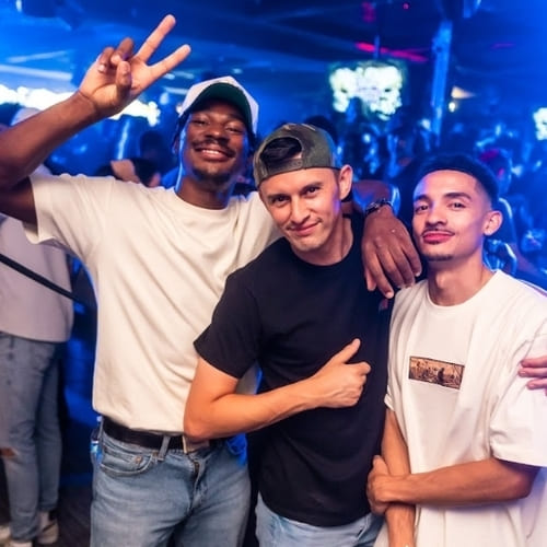 Three men posing and smiling at a nightclub, one flashing a peace sign while others have their arms around each other.