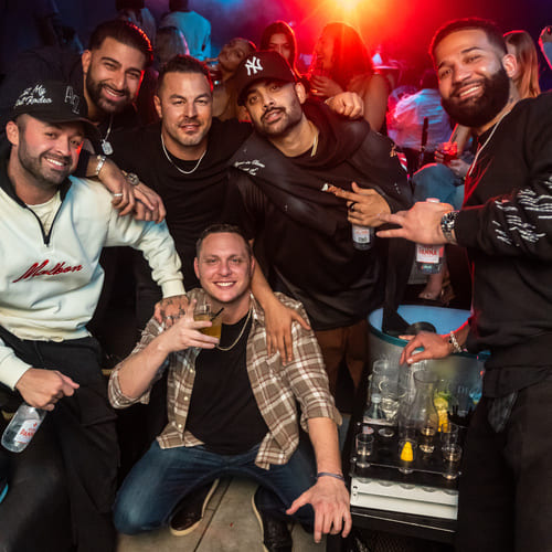 Group of men posing and smiling at a nightclub VIP table with drinks and red lighting in the background.