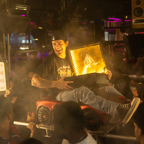 Man holding glowing golden Ace of Spades champagne sign while seated on a red cart in a crowded nightclub with smoke and lights
