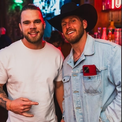 Two men pose together at a nightlife venue, one wearing a white shirt and the other in a cowboy hat and denim jacket, both smiling at the camera.