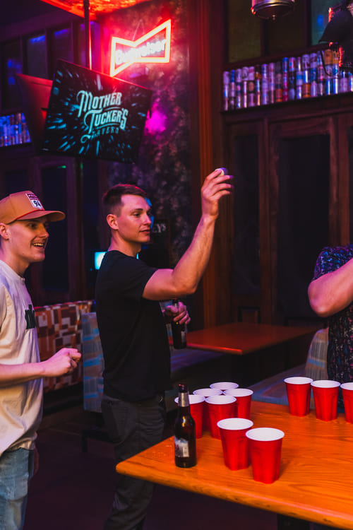 Three men play beer pong with red cups and a beer bottle on a wooden table under neon lighting in a bar.