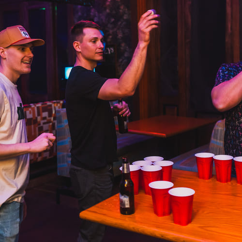 Three men play beer pong with red cups and a beer bottle on a wooden table under neon lighting in a bar.