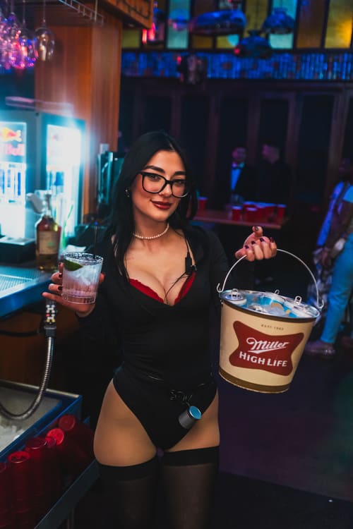 Bartender in a black bodysuit and glasses holding a Miller High Life beer bucket and cocktail behind a bar at a nightlife venue.