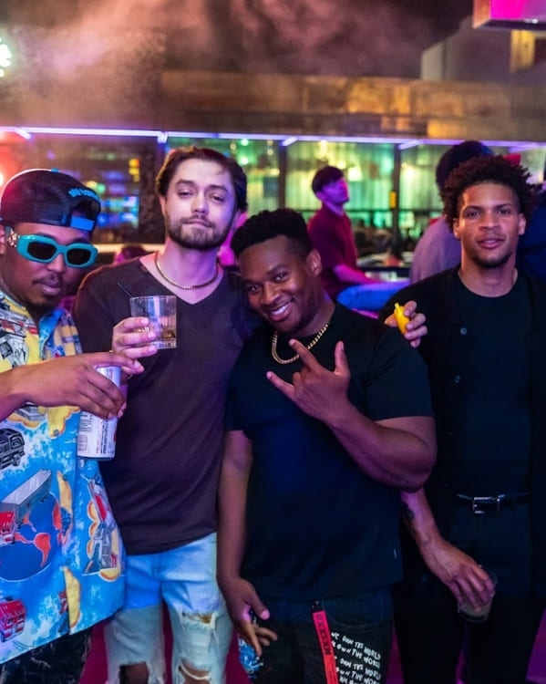 Four men posing for a photo at a nightclub, holding drinks and smiling under colorful lights.