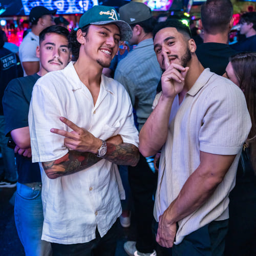 Three men pose for a photo at a nightclub, smiling and gesturing with peace signs, surrounded by colorful lights and a crowd.