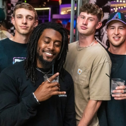 Four men smiling and holding drinks while posing for a photo at a lively nightlife venue.
