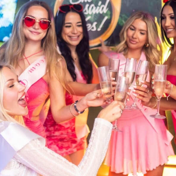 Group of women dressed in pink outfits clinking champagne glasses in a celebratory toast at a bachelorette party.