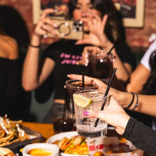 Close-up of several women raising cocktails for a toast at a table with nachos, fries, and dipping sauces, one woman taking a photo.