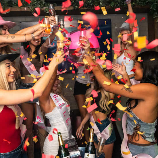 Group of women in festive outfits raising champagne glasses with confetti falling during a bachelorette party celebration.