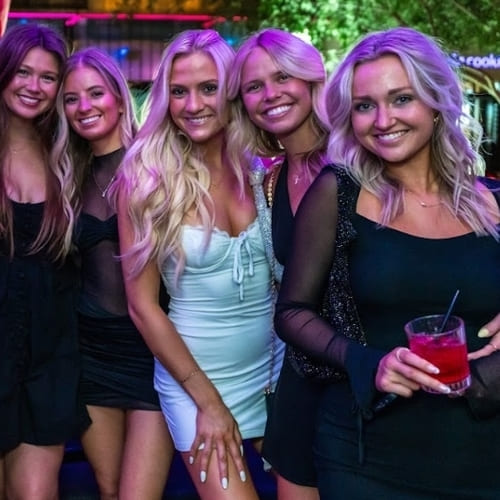 Group of five women in stylish black and white dresses posing and smiling during a night out with vibrant pink and purple lighting.