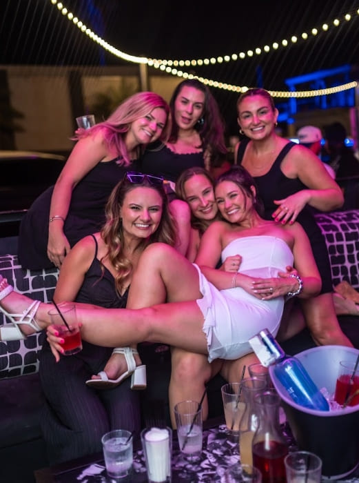 Group of six women dressed in black and white party outfits celebrating in a nightclub VIP booth with drinks and smiling for a photo