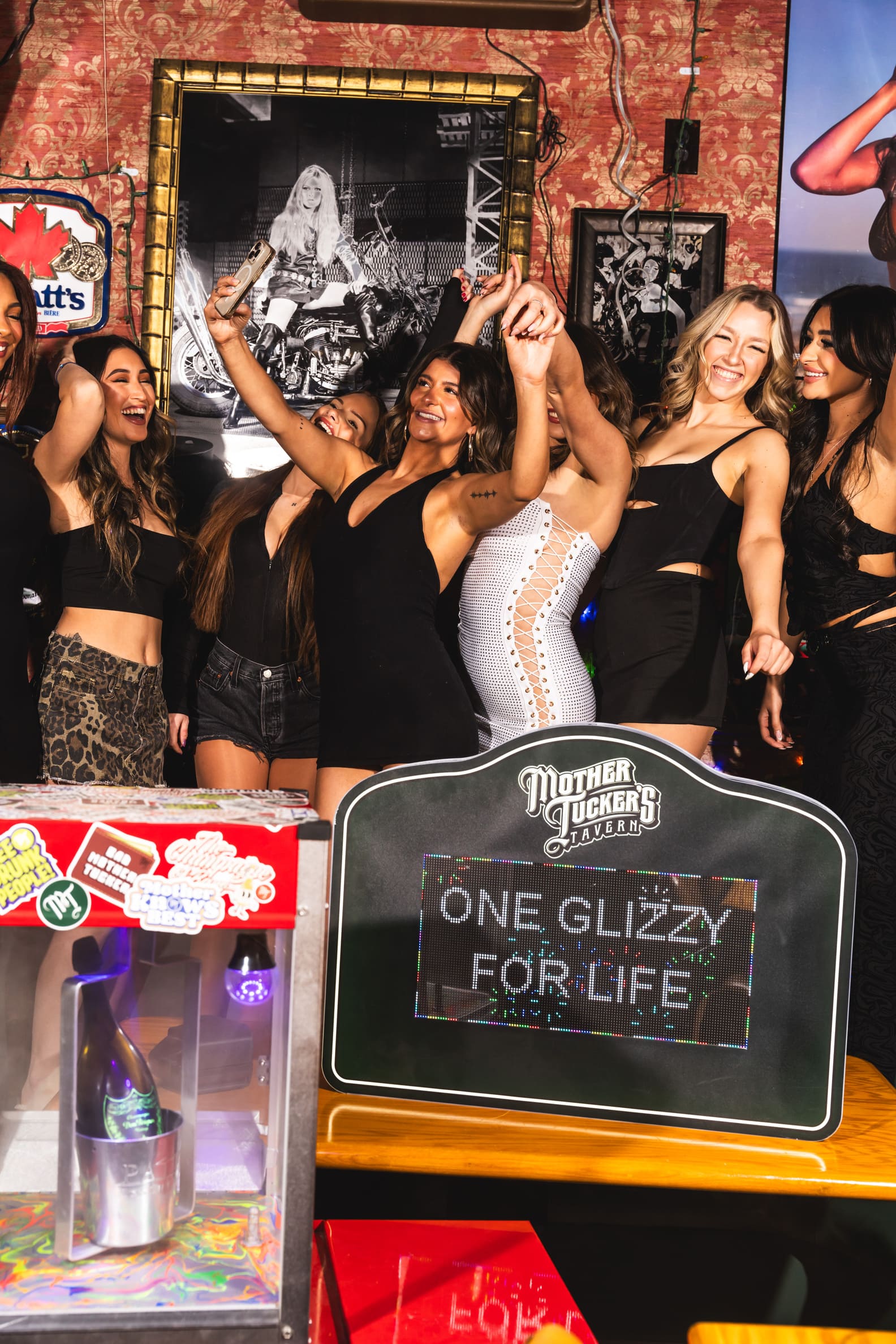 Group of women in black and white outfits celebrating indoors, standing behind a humorous sign that reads “ONE GLIZZY FOR LIFE.”
