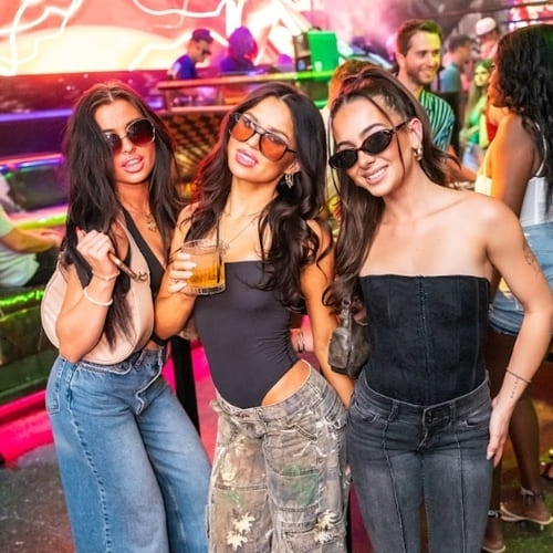 Three women in sunglasses and stylish outfits posing together at a vibrant nightclub