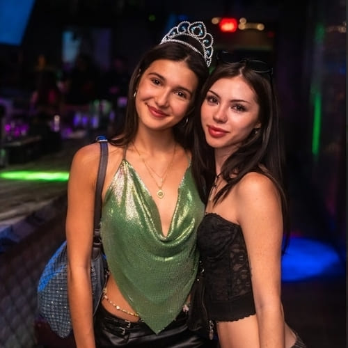 Two young women posing together at a nightclub; one wears a green sparkly halter top and tiara, the other wears a black lace corset top, both smiling.