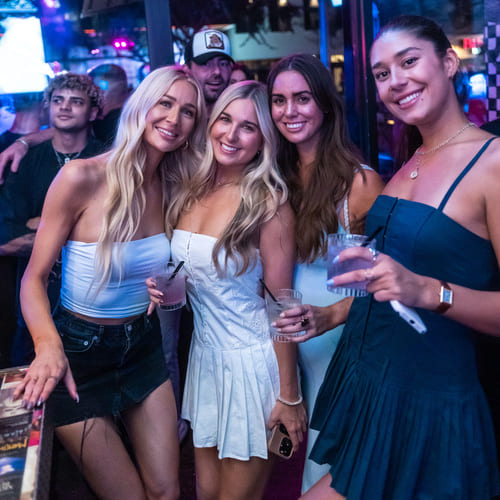 Four women smiling and holding drinks in a vibrant nightclub atmosphere.