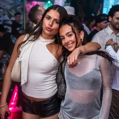 Two women smiling and posing together at a nightclub, holding drinks and standing close under colorful lights.