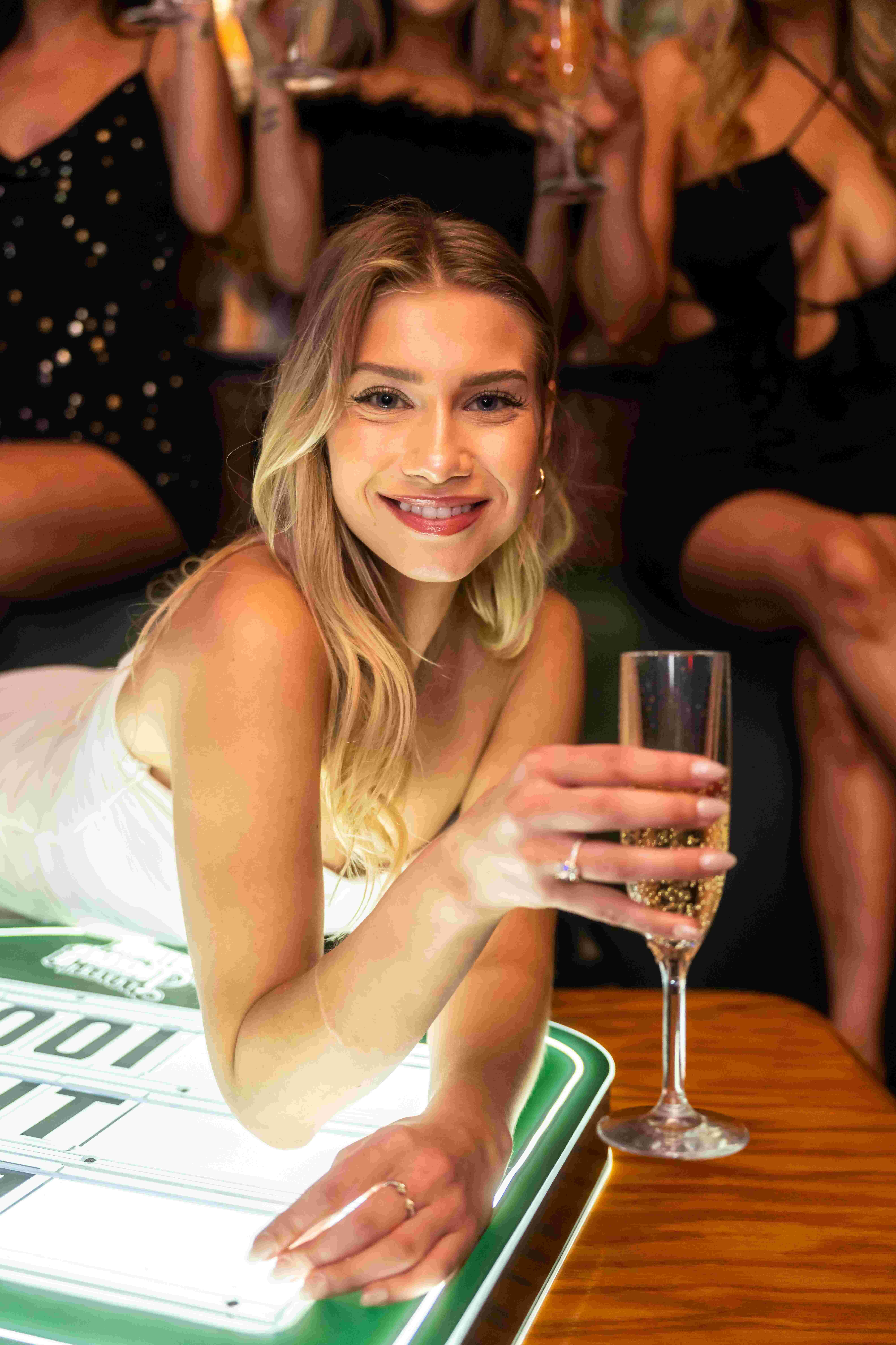 Smiling woman celebrating with champagne at a bachelorette party, surrounded by friends in glamorous outfits. A vibrant nightlife moment full of joy, style, and festive energy