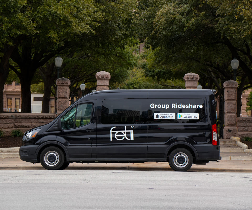 Black Fetii group rideshare van parked on a city street with stone pillars and trees in the background.